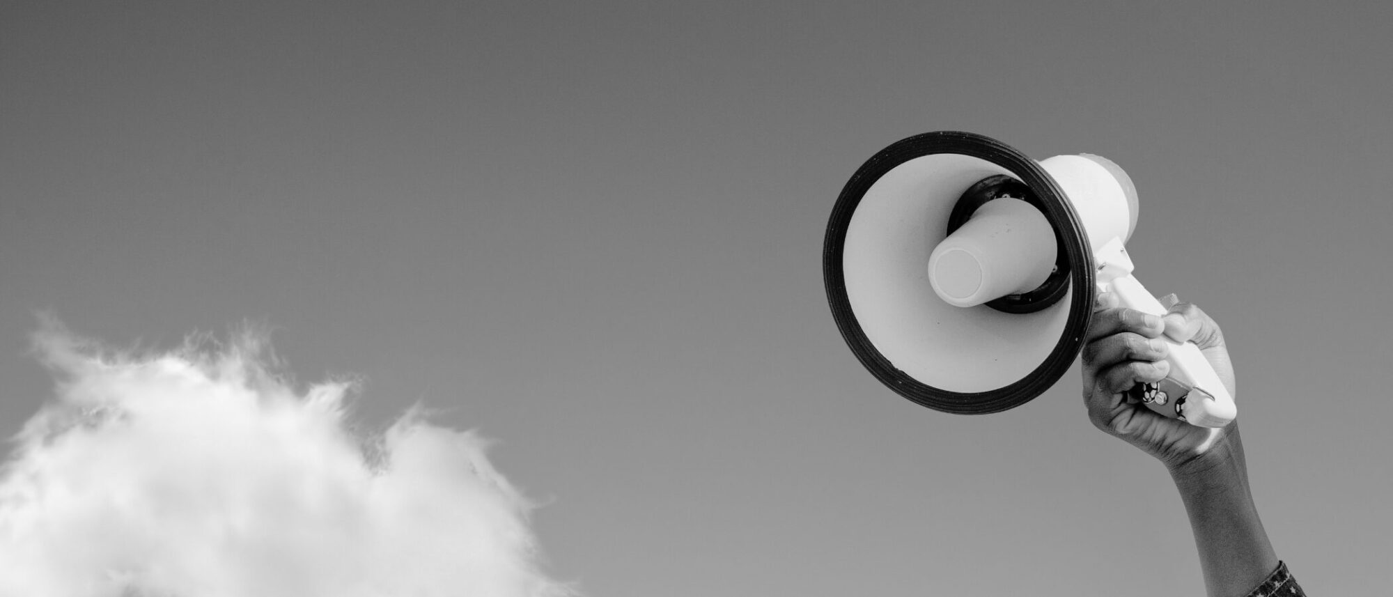 A hand holding a megaphone is raised against a clear sky with one cloud visible. The image is in black and white.