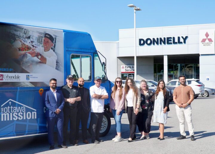 A group of Donnelly employees and Chef Ric stand in front of a blue Ottawa Mission truck in front of a Donnelly car dealership. The atmosphere is friendly and sunny.