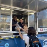 A woman with a backpack receives a bag of food from two smiling food truck chefs.