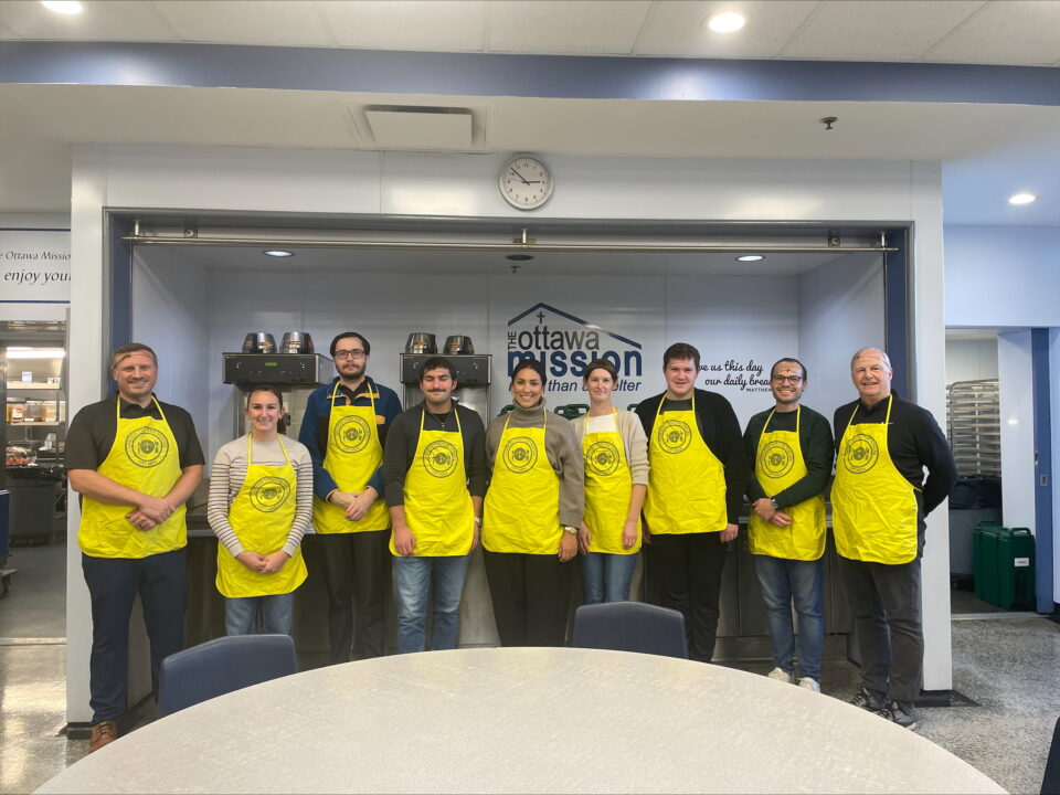 A group of nine people in yellow aprons stand smiling in a kitchen with "Ottawa Mission" signage. Stainless steel equipment is visible behind them.