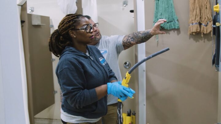 A young black woman, student of the MSTP, is wearing personal protective equipment and paying attention to the instructor pointing at a drywall.