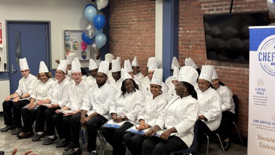A group of 25 graduates sit at their ceremony, wearing white chef attire.