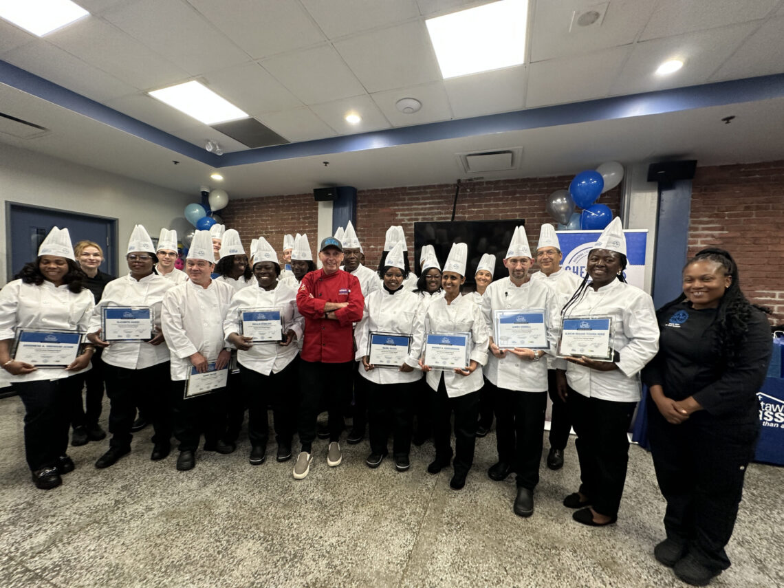 A group of 25 graduates pose, smiling in chef uniforms and holding diplomas.