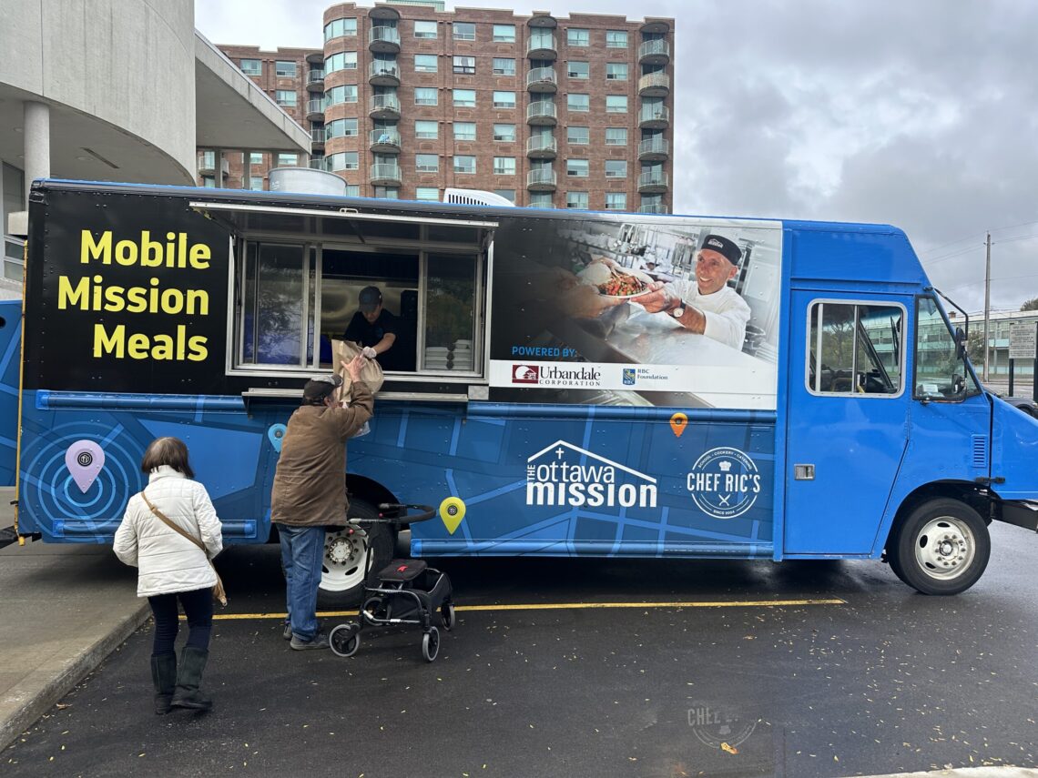 A man hands a brown bag of groceries from out the window of a food truck which reads "Mobile Mission Meals"