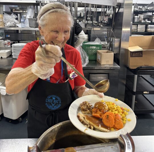 A smiling older woman pours gravy on top of a turkey dinner.