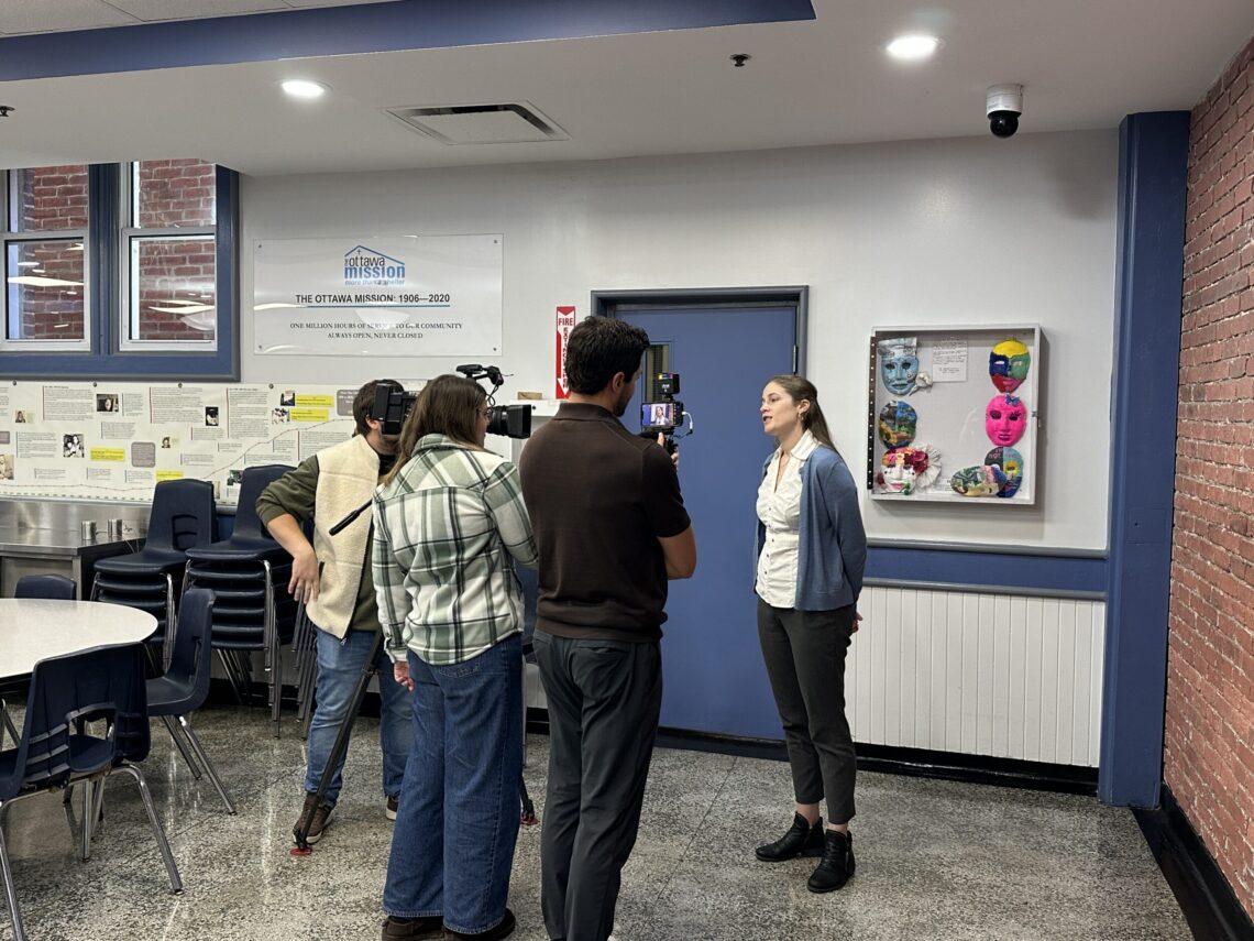 A woman speaks to reporters inside a well lit dining room at The Ottawa Mission.