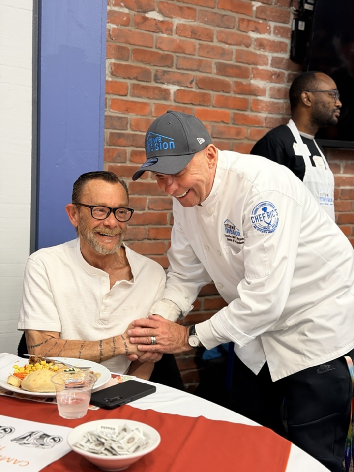 A man in a chef uniform smiles and holds the hand of another man sitting at a table with a plate of turkey and fixings in front of him.